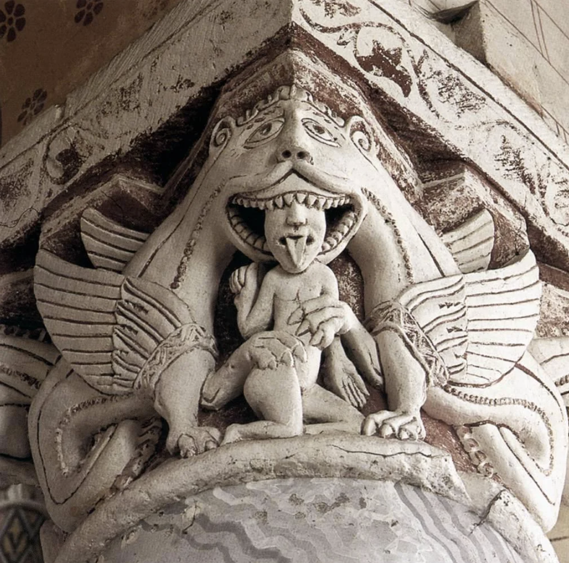 Steinrelief: geflügeltes Monster mit offenem Maul, das eine kleine menschliche Figur mit herausgestreckter Zunge hält.